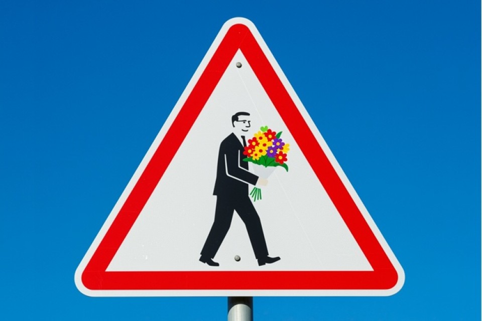 Tegen blauwe achtergrond een wit driehoekig verkeersbord met rode rand, middenin een zakenman met een bos bloemen in zijn hand