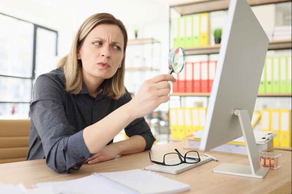 Vrouw kijkt sceptisch naar computerscherm terwijl ze een vergrootglas vasthoudt aan bureau met bril en notitieblok