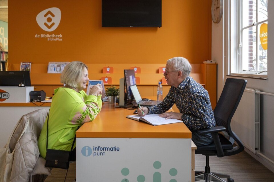 Medewerker en bezoeker zitten aan een oranje balie in de Bibliotheek BiblioPlus, met computers en informatiemateriaal.