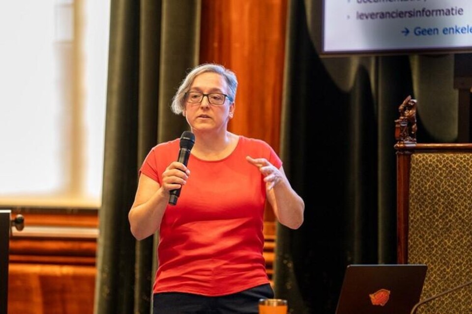 Vrouw met bril en rood shirt spreekt met microfoon op podium, met scherm en laptop op de achtergrond.