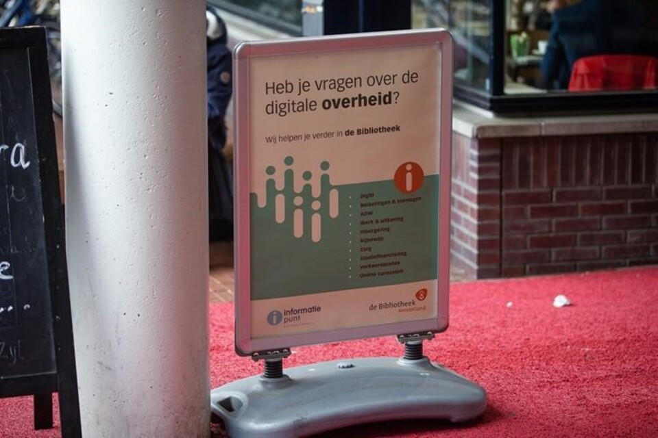 Informatiebord met tekst over digitale overheid en hulp in de bibliotheek, geplaatst op een grijze standaard op rode vloer.
