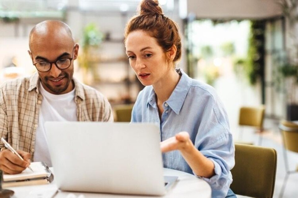 Man en vrouw zitten aan tafel, kijken naar laptop, man maakt aantekeningen, lichte kantooromgeving.
