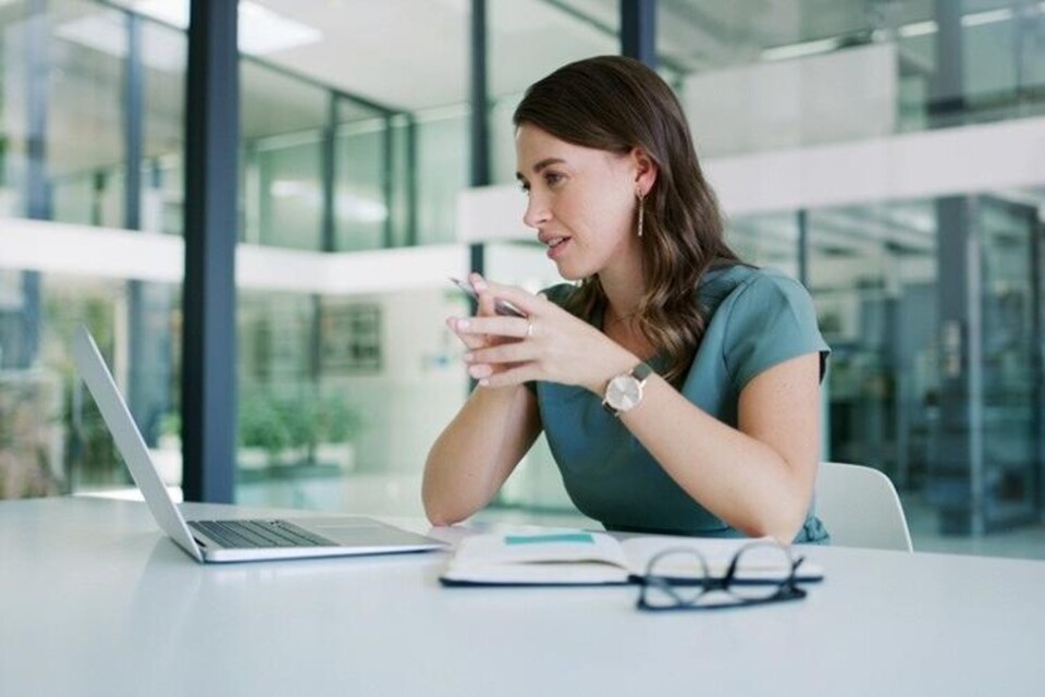 Vrouw zittend aan bureau met laptop, notitieboek en bril, werkend in een lichte kantoorruimte.