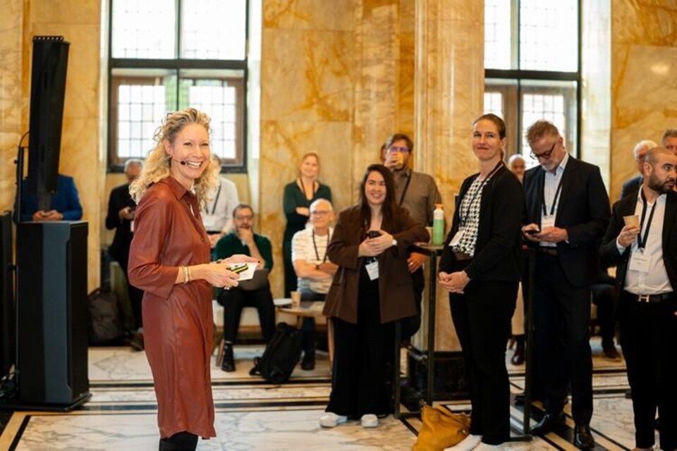 Lachende vrouw in roodbruine jurk met microfoon voor een zaal met toehoorders.