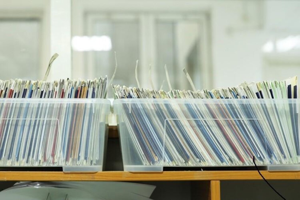 Plastic bakken gevuld met papieren dossiers en mappen, staand op een houten plank in een archiefruimte.