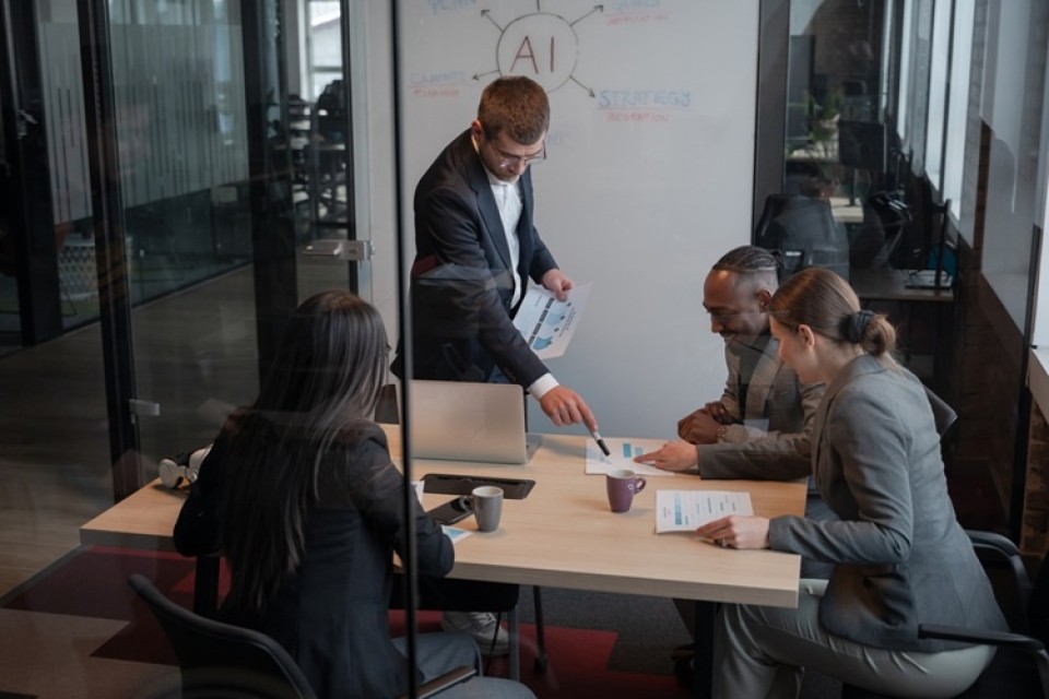 Twee mannen en twee vrouwen zitten rondom een vergadertafel. Op de achtergrond een whiteboard met AI erop geschreven
